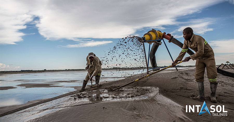 Two men dressed for work are engaged in pumping tailings into a tailings storage facility using a mechanical pump.