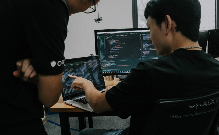 A man writing code on a laptop while another person observes